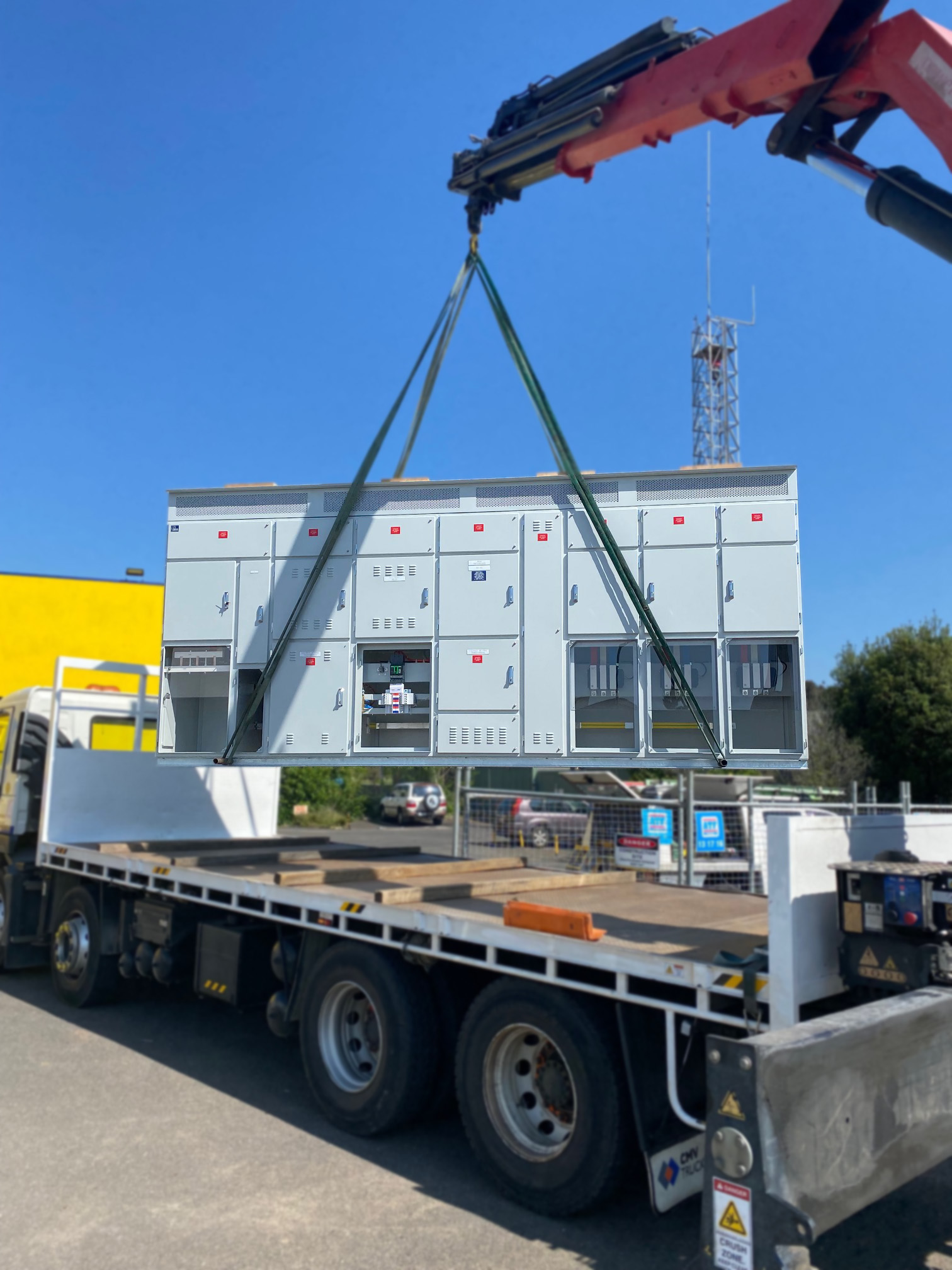 Electrical switchboard being hoisted by a crane truck, secured with rigging straps during transport setup at an industrial site